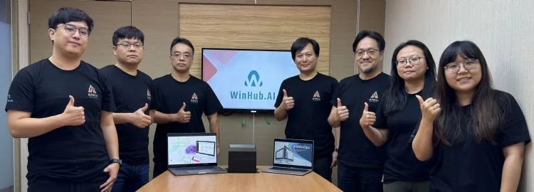 The AIWin team in company T-shirts giving a thumbs up, with WinHub.AI on the screen behind them and AIWinOps on the laptops